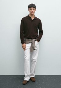 Young man wearing a dark brown long-sleeve polo, white pants, brown shoes, and a gray sweater tied around his waist, standing against a gray wall.
