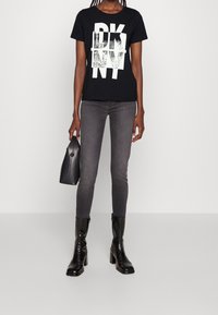 Svart bomulls t-shirt med vit "DKNY" grafik, kombinerad med grå slim-fit jeans och svarta ankelstövlar, medan hon håller en liten svart handväska.