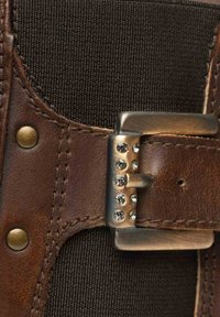Cintura in pelle marrone con fibbia in metallo decorata con strass trasparenti, attaccata a un tessuto scuro testurizzato con rivetti in ottone e cuciture dettagliate.