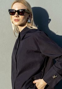 Blondynka w czarnych okularach przeciwsłonecznych, z dużymi okrągłymi kolczykami i czarną koszulą z detalem rozgwiazdy na mankiecie, stoi na tle szarej ściany.