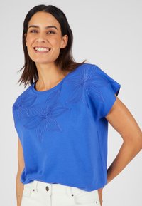 T-shirt in cotone blu con ricami floreali sulla parte anteriore, scollatura rotonda e maniche corte, abbinato a pantaloni bianchi.