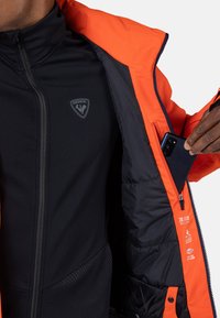 Svart isolerad jacka med orange detaljer, med en ficka med dragkedja. Tyg har en slät textur och visar vatten- och vindavvisande egenskaper.