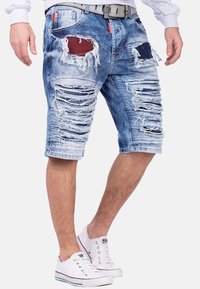 Shorts en denim bleu clair avec des zones déchirées, des bords effilochés et des détails en patchwork. Comprend un design à cinq poches et des accents en métal.