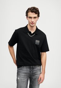 Ung man iklädd en svart Hugo-pikétröja, med kedja runt halsen och blekta grå jeans, stående mot en ljus enfärgad bakgrund.