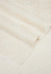 Vaalea beige kudottu viltti, jossa on hillitty timanttikuvio ja fringe-reunat, esittelee sileää tekstuuria ja pehmeää viimeistelyä.