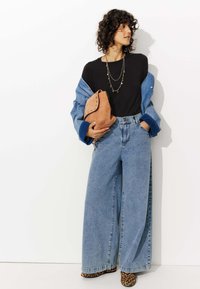Pantalon en denim bleu à jambes larges, haut noir et veste en denim clair. Tenant un sac en daim beige, orné de clous en argent. Chaussures à imprimé léopard.