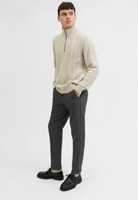 Bézs színű, kábelmintás pulóver magas gallérral, sötétszürke, szabott nadrággal és fekete cipővel párosítva. A pulóver részletes textúrával rendelkezik.