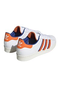 Bílé kožené tenisky s oranžovými a modrými akcenty, texturovanými pruhy a gumovou podešví. Mají klasický nízký střih a zavazování na tkaničky.