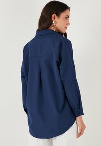 Camicia a maniche lunghe blu navy in tessuto leggero, con una piega sul retro, colletto a punta e dettagli dei polsini con bottoni. Texture liscia.