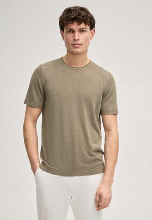 Junger Mann mit lockigem Haar, trägt einen olivgrünen Kurzarm-Pullover und weiße Hosen, steht mit einer Hand in der Tasche.