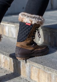 Bota de invierno de color marrón y negro con parte superior acolchada, cuello de piel sintética, cierre de cordones en la parte delantera y suela de goma resistente para mayor tracción en las superficies.
