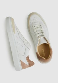 Baltas ādas kedas ar perforētu dizainu, bēša zamšādas akcentiem un baltu gumijas zoli. Iespējas ietver noapaļotu purngalu un saites priekšpusi.