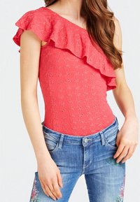 Body en corail à une épaule avec un col à volants, orné de motifs floraux brodés. Associé à un jean en denim présentant des accents floraux.