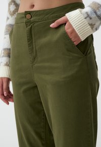 Pantaloni in cotone verde oliva con tasche laterali, chiusura a bottone e texture morbida. Presentano una vita aderente e una forma delle gambe rilassata.