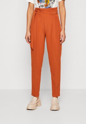 Hoch taillierte, rostorange, schmal zulaufende Hose mit Stoffgürtel, Vorderfalten, getragen mit beigen, klobigen Loafers und einem Top mit floralem Totenkopf-Print.