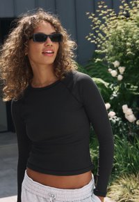 Zwart geribd shirt met lange mouwen en een strakke pasvorm, gecombineerd met grijze joggingbroek met trekkoord. Omringd door groen en bloemen.