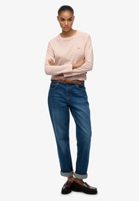 Helles rosa Langarmshirt mit Rundhalsausschnitt, kombiniert mit blauen High-Waist-Jeans und einem geflochtenen braunen Gürtel. Braune Slipper.