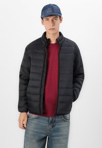 Black padded jacket with a zip front and stand collar, paired with a red sweater. The model wears a blue cap and light blue jeans.