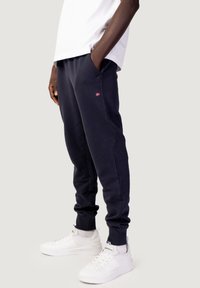 Persona che indossa pantaloni da jogging blu navy con un piccolo logo a bandiera sulla coscia, calzini bianchi, sneakers bianche e una camicia bianca, mani in tasca.