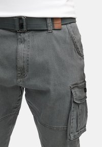 Pantalon cargo gris avec poche latérale, ceinture noire tissée avec boucle métallique, et chemise blanche rentrée à la taille.