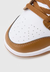 Brązowo-biały sneaker z gładką skórzaną cholewką, perforowanym przodem, teksturowanymi brązowymi sznurowadłami i kontrastową gumową podeszwą.