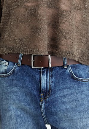 Bruine leren riem met zilveren metalen gesp, gedragen over blauwe denim jeans met zichtbare stiksels en een vervaagde textuur. De trui heeft een gestructureerd patroon.