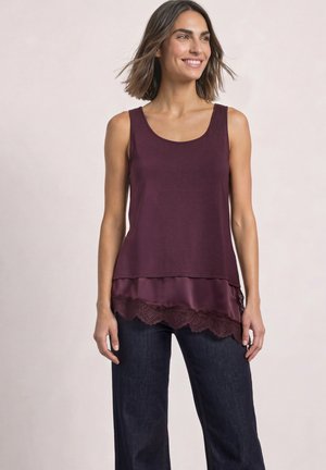 Femme souriante portant un haut sans manches violet foncé avec un ourlet en dentelle et un jean bleu foncé, debout devant un fond clair uni.