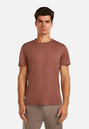 Ung mand, der står med front mod kameraet iført en ensfarvet brun t-shirt med korte ærmer og lysebrune bukser mod en hvid baggrund.