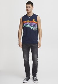 Maglietta senza maniche blu navy con un design a onde multicolore sul petto, abbinata a jeans scuri e sneakers alte nere.