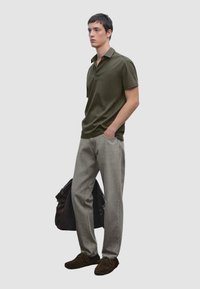 Olivově zelené polo tričko, světle šedé džíny a hnědé semišové boty. Model drží tmavě hnědou tašku a stojí před neutrálním pozadím.