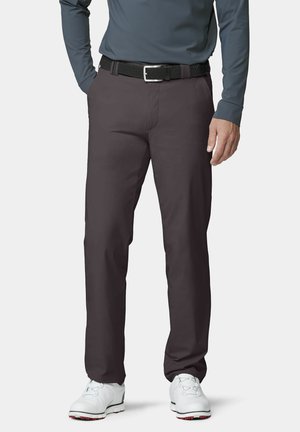 Mann trägt dunkelgraue Hose, schwarzen Gürtel, graues Langarmshirt und weiße Golfschuhe, steht mit einer Hand in der Tasche.