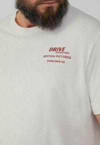 Jasno beżowy t-shirt z okrągłym dekoltem, wykonany z miękkiego materiału, z nadrukowanym na czerwono tekstem: „DRIVE STUNT UNIT MOTION PICTURES 9362-309-13.”