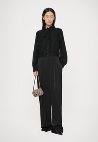 Blouse noire à sequins avec un nœud papillon au col, associée à un pantalon noir à jambes larges. Le mannequin tient un petit sac à main gris. Texture lisse.