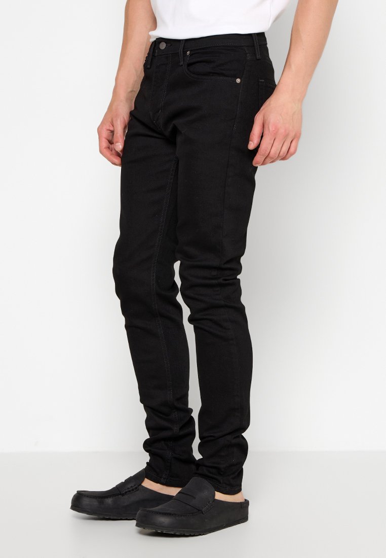 Jeans neri slim-fit realizzati in denim con una superficie liscia. Caratteristiche includono un design a cinque tasche e cuciture a contrasto. Indossati con scarpe slip-on nere.