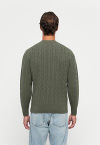 Mann som står med ryggen til kameraet iført en olivengrønn kabelstrikket genser og lyseblå jeans mot en ensfarget hvit bakgrunn.