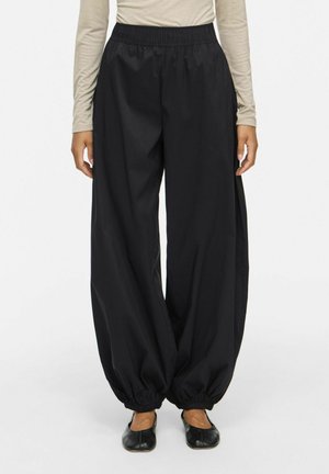 Personne portant un pantalon noir ample à taille élastique avec des manches élastiquées froncées et des chaussures plates noires, assorti à un haut beige à manches longues.