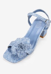 Sandalia de tacón alto de denim azul con punta cuadrada, acento floral, tira en el tobillo y tejido texturizado. El tacón cuadrado proporciona estabilidad.