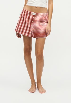 Femme portant un short à carreaux rouges et blancs et un haut blanc sans manches, debout pieds nus sur un fond blanc uni.