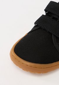 Baskets en toile noire avec des bandes velcro, une semelle en caoutchouc naturel et une tige texturée. Vue rapprochée montrant la forme de la chaussure et les détails de la couture.