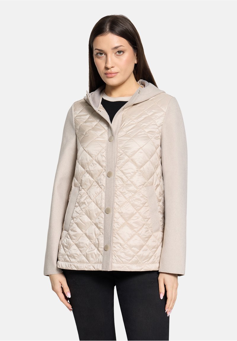 Femme portant une veste matelassée beige avec boutons-pression, manches longues et poches latérales, debout devant un fond clair uni.