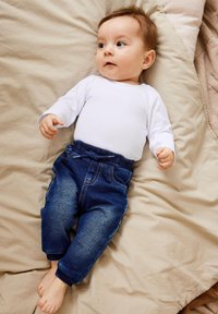 Witte lange mouwen bodysuit en donkerblauwe denim broek met elastische tailleband, voorzien van een trekkoord en lichte wassing accenten, gelegd op beige stof.
