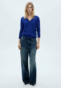 Maglione blu a collo a V con maniche lunghe abbinato a jeans in denim scuro a vita alta e gamba larga. Texture morbida con leggeri sbiadimenti sui jeans.