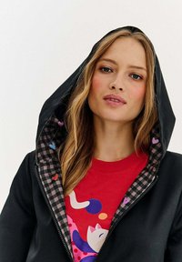 Jauna sieviete ar blondiem matiem valkā melnu pārbaudi rīkotu jaku virs sarkanās kreklas ar krāsainu kaķi un abstrahētām formām.