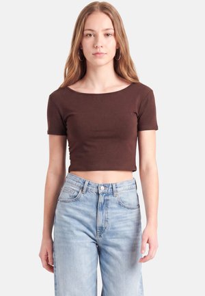Ung kvinde med langt lysbrunt hår, iført øreringe i ringe, en brun croppet korteærmet top og lyseblå højtaljede jeans, stående.