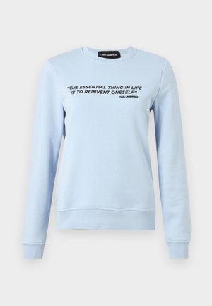 Sudadera azul claro hecha de tejido suave, con mangas largas y cuello redondo. En la parte frontal, hay un texto negro que cita a Karl Lagerfeld.