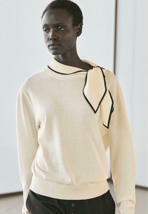 Femme aux cheveux courts portant un pull crème avec un détail écharpe sur une épaule et un pantalon noir, regardant sur le côté à l'intérieur.