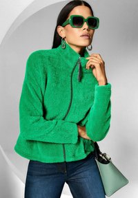 Frau trägt eine leuchtend grüne, flauschige Zip-Jacke, große grüne Sonnenbrille, silberne Ohrringe, dunkle Jeans und hält eine hellgrüne Handtasche.