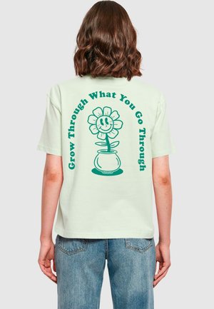 Hellgrünes, übergroßes Baumwoll-T-Shirt mit einem Grafiker eines lächelnden Blumenmotivs in einem Topf und dem Text "Wachse durch das, was du durchmachst."