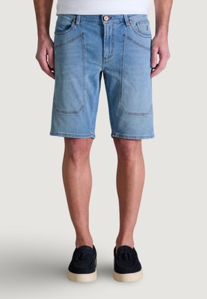 Hombre que lleva pantalones cortos de mezclilla azul claro con grandes bolsillos delanteros y zapatos negros sin cordones con borlas, de pie contra un fondo liso.