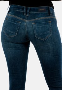 Jeans de denim azul oscuro con una forma ajustada, que presentan bolsillos traseros y detalles de costura sutiles. Etiqueta de la marca visible en la cinturilla.
